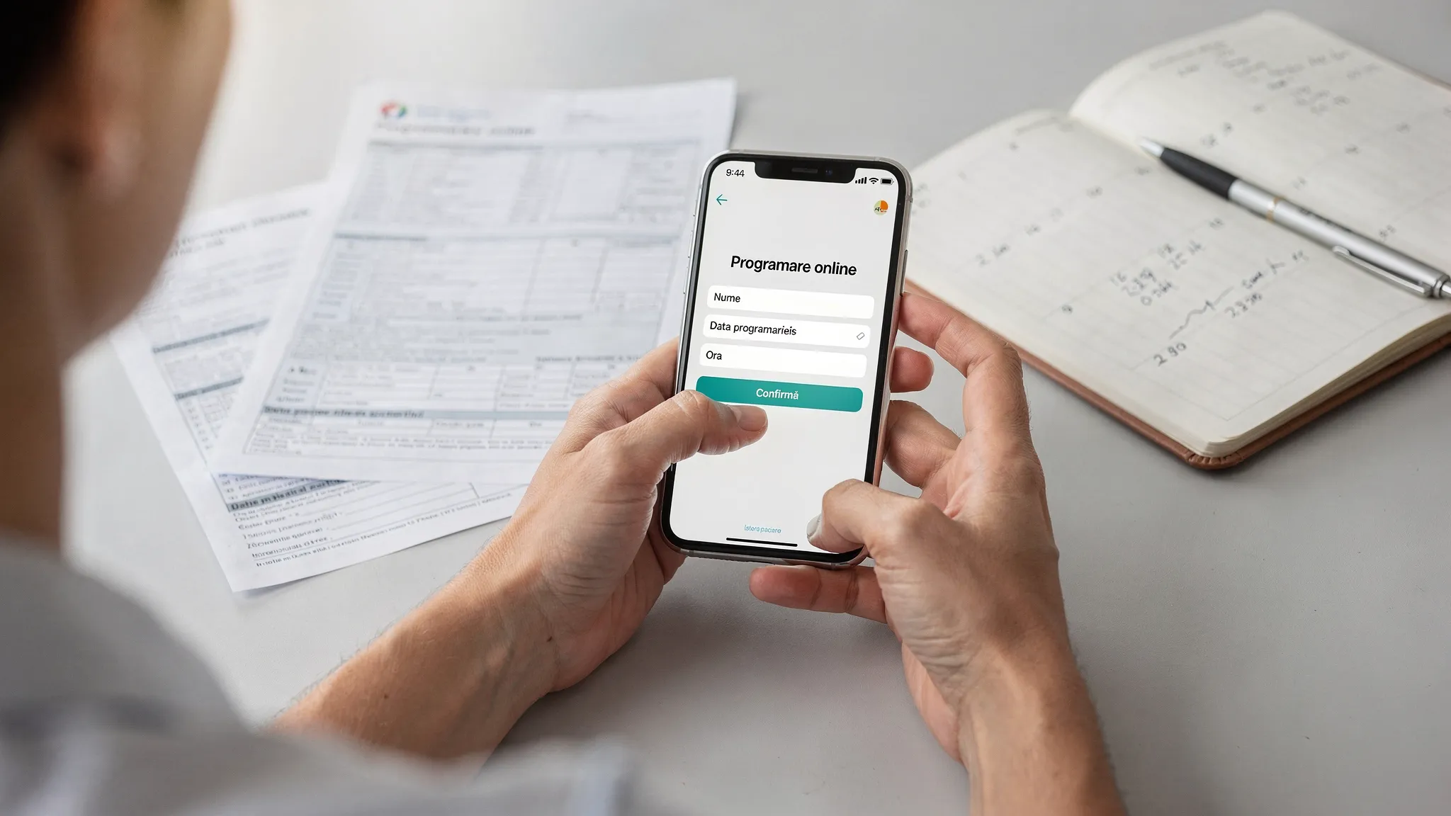 O persoană completează un formular de programare online pe telefon, lângă ea se află o agendă deschisă și câteva documente medicale, sugerând organizare înainte de consultație.