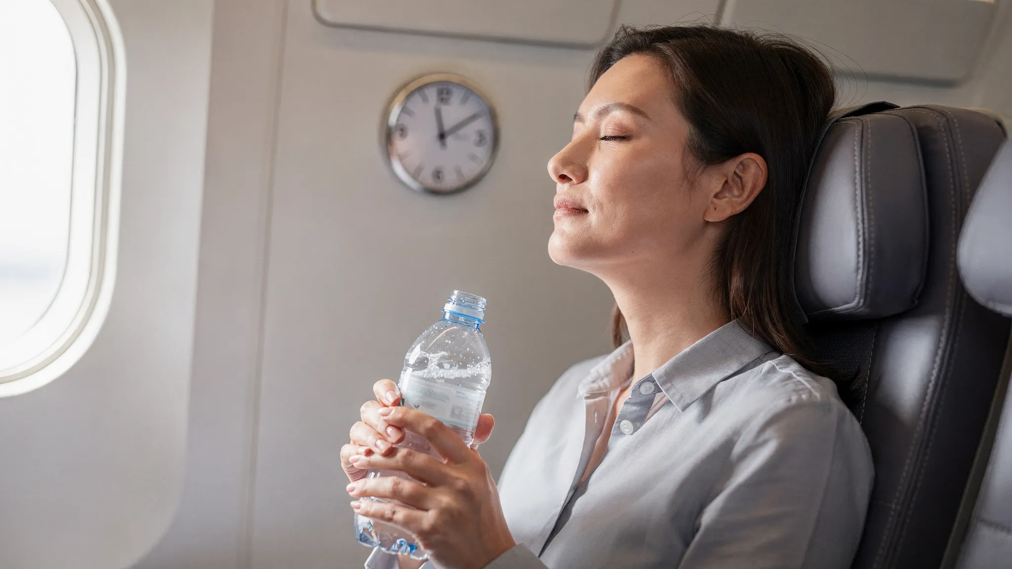 O persoană într-un scaun de avion, cu capul sprijinit corect pe tetieră fără să apese fața în pernă, având în mână o sticlă de apă; în imagine se vede discret un ceas care sugerează respectarea intervalului de câteva ore după o procedură estetică.