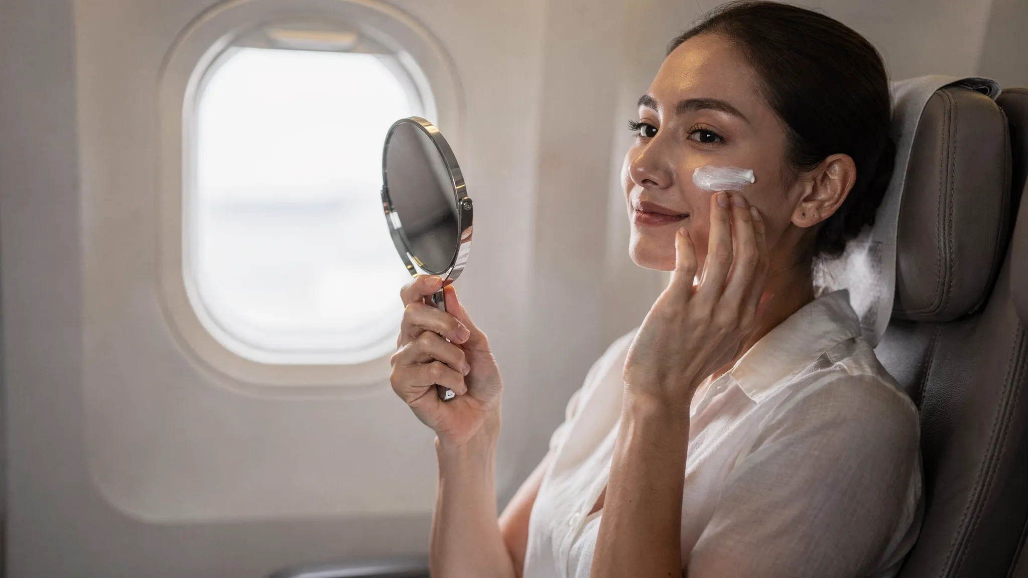 O persoană stă pe scaun într-o cabină de avion, cu o oglindă mică în mână și un gest delicat de aplicare a unei creme hidratante, sugerând îngrijire blândă după un tratament estetic; în fundal se văd hubloul și tetierele, fără ecrane vizibile.